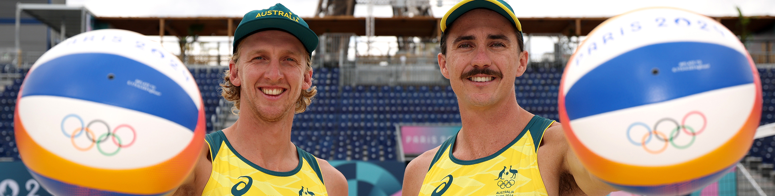 Zachery Schubert and Thomas Hodges hold beach volleyballs with Olympic rings in front of an empty stand
